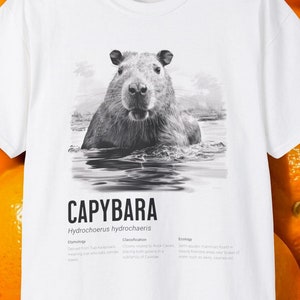 May include: A white t-shirt featuring a black and white illustration of a capybara swimming in water. The text "CAPYBARA Hydrochoerus hydrochaeris" is printed below the image. The text "Etymology, Classification, Ecology" is printed below the text "CAPYBARA".