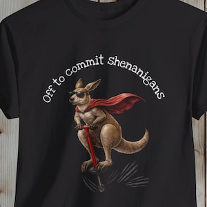 Op de afbeelding: Zwart t-shirt met een afbeelding van een kangoeroe met een zonnebril en een rode cape, rijdend op een pogo-stick. De tekst "Off to commit shenanigans" staat boven de kangoeroe. Het shirt wordt getoond tegen een houten achtergrond.