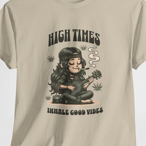 May include: A beige t-shirt featuring a cartoon illustration of a woman playing a guitar and smoking. The text "HIGH TIMES" is above the illustration and "INHALE GOOD VIBES" is below.