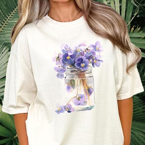 Op de afbeelding: Een gebroken wit t-shirt met korte mouwen en een grafische print van een doorzichtige glazen pot gevuld met water en een boeket paarse en lichtblauwe viooltjes, met een paar losse bloemblaadjes onderaan de pot.