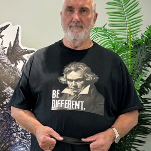 Pu&ograve; includere: Un uomo indossa una maglietta nera con un ritratto di un uomo e il testo "BE DIFFERENT." Indossa una cintura nera e un orologio al polso sinistro.