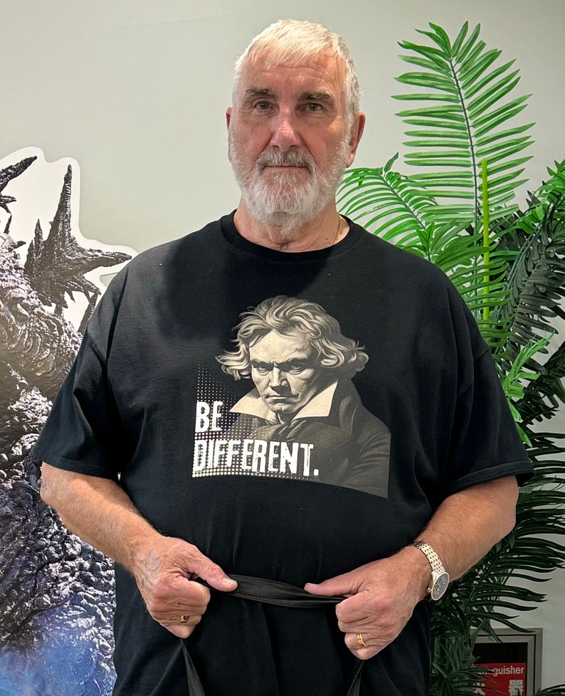 Pu&ograve; includere: Un uomo indossa una maglietta nera con un ritratto di un uomo e il testo "BE DIFFERENT." Indossa una cintura nera e un orologio al polso sinistro.