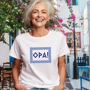 May include: White t-shirt with a blue and white patterned frame around the word "OPA!" in blue.