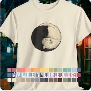 以下が含まれることがあります： 胸元に円形のグラフィックがプリントされたクリーム色の半袖Tシャツ。グラフィックには、陰陽の形に丸まった黒猫と白猫が描かれています。グラフィックの下には「AVAILABLE IN MULTIPLE COLORS」というテキストが記載されています。