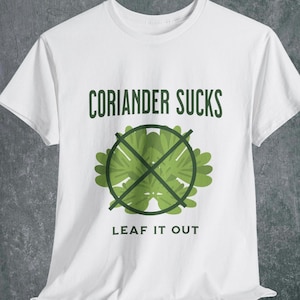 Puede incluir: Camiseta blanca con el texto "CORIANDER SUCKS" en verde oscuro sobre un gráfico verde de hojas de cilantro tachadas. Debajo del gráfico, el texto "LEAF IT OUT" está impreso en verde oscuro.