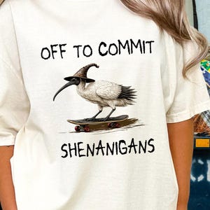 Op de afbeelding: Een gebroken wit t-shirt met een afbeelding van een ibisvogel die een puntige bruine hoed draagt terwijl hij op een skateboard rijdt. De tekst boven de vogel luidt OFF TO COMMIT en de tekst eronder SHENANIGANS in een zwart, handgetekend lettertype.