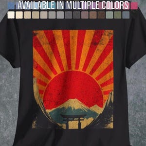 Op de afbeelding: Een zwart t-shirt met een grafische print van een rode zon met oranje stralen die naar buiten stralen, geplaatst boven een bergtop en een torii-poort. Het ontwerp heeft een verweerde textuur. De tekst "AVAILABLE IN MULTIPLE COLORS" is zichtbaar.
