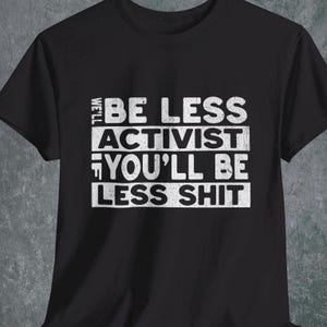 Op de afbeelding: Zwart T-shirt met witte tekst: "BE LESS ACTIVIST YOU'LL BE LESS SHIT". De tekst is in drie regels gestapeld. Het T-shirt is gemaakt van een zacht materiaal.