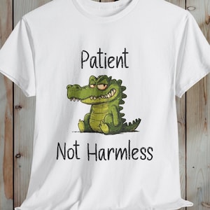 May include: White t-shirt featuring a cartoon alligator with the text "Patient" above and "Not Harmless" below. The alligator is green with a grumpy expression. The shirt is a casual wear item.