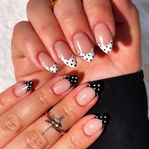 Pode incluir: Close-up de unhas cuidadas com um design de ponta francesa. As pontas alternam entre branco com bolinhas pretas e preto com bolinhas brancas. As unhas são ovais e têm um acabamento brilhante. Anéis dourados adornam os dedos.