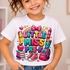 Puede incluir: Una camiseta blanca de manga corta con un estampado gráfico colorido con el texto LITTLE MISS PRE-K. El diseño incluye un lápiz, una manzana, una pila de libros, zapatillas, una mochila y un arcoíris, decorados con corazones y estrellas.