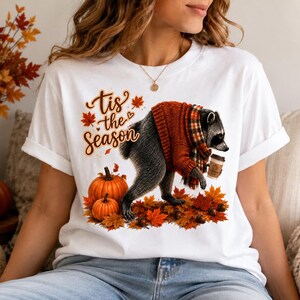 May include: A white t-shirt featuring a graphic of a raccoon wearing an orange jumper and a plaid scarf, holding a coffee cup. The design includes orange pumpkins, scattered autumn leaves, and the text "tis the season" in brown script lettering.