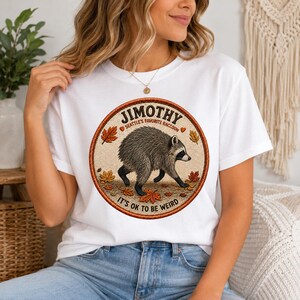 以下が含まれることがあります： 白いTシャツで、オレンジや黄色の秋の葉の中を歩く茶色のラクーンが描かれた円形のグラフィックが特徴です。ラクーンのイラストの周りには、濃い茶色の文字で「JIMOTHY SEATTLE'S FAVORITE RACCOON」および「IT'S OK TO BE WEIRD」と書かれています。