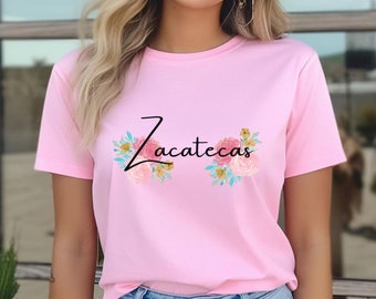 May include: A light pink t-shirt featuring the word "Zacatecas" in black script, adorned with floral arrangements in pink, yellow, and blue. The shirt is a crew neck style.