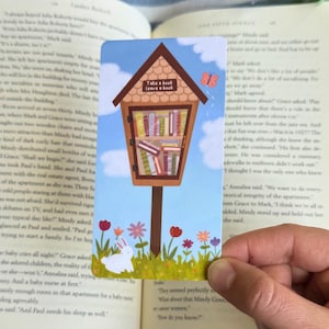 May include: A bookmark depicting a small wooden library with the text "Take a book, leave a book." The design includes a shelf of books, a white rabbit, flowers, and a butterfly. The bookmark is held over an open book.