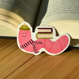 May include: A pink worm sticker with a green hat, striped scarf, and two books. The worm has a smiling face and is lying down. The sticker is on a wooden surface with an open book in the background.