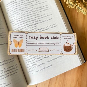 May include: A rectangular bookmark with the text "cozy book club" and illustrations of a butterfly, a coffee cup, and a book. The bookmark is on top of an open book.