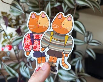 Puede incluir: Dos gatos tabby naranja con suéteres y mochilas. El gato de la izquierda lleva un suéter floral rosa y el gato de la derecha lleva un suéter a rayas.