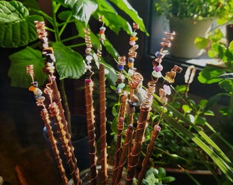 May include: A collection of copper-wrapped wands adorned with colorful gemstones. The wands are arranged in a glass container, with the copper wire spiraling around the wooden shafts. The gemstones include various colors and shapes, adding a touch of natural beauty.
