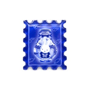 Pu&ograve; includere: Un francobollo decorativo blu con un personaggio di coniglio bianco che indossa una camicia blu con un disegno a farfalla. Il coniglio &egrave; ambientato contro un vaso blu con un disegno di drago. Il francobollo ha un bordo smerlato e uno sfondo bianco.