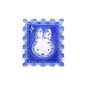 Pu&ograve; includere: Un adesivo blu a forma di francobollo con un design a coniglietto bianco con vortici decorativi e una stella. L'adesivo ha un bordo a pois e un bordo smerlato, creando un'estetica stravagante.