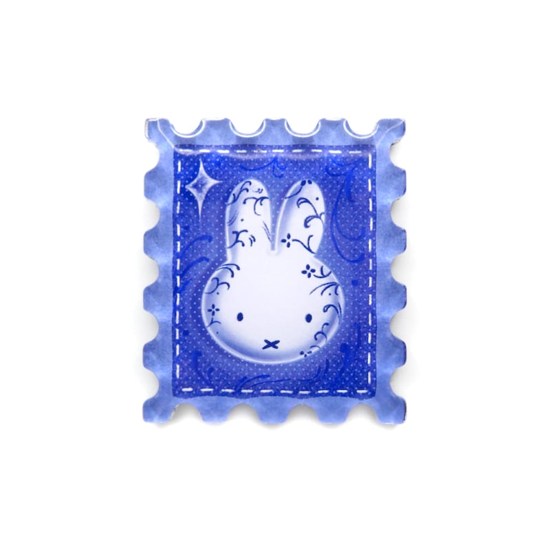 Pu&ograve; includere: Un adesivo blu a forma di francobollo con un design a coniglietto bianco con vortici decorativi e una stella. L'adesivo ha un bordo a pois e un bordo smerlato, creando un'estetica stravagante.