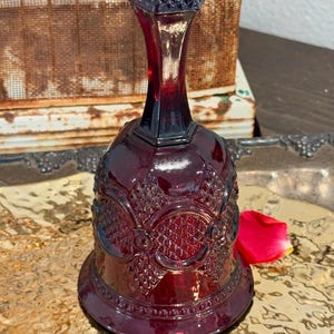 Puede incluir: Una campana de cristal rojo intenso con un mango facetado y un cuerpo decorado con un patrón de diamantes prensados y motivos circulares. La campana tiene una base acampanada con un borde de cuentas y una superficie lisa y reflectante.