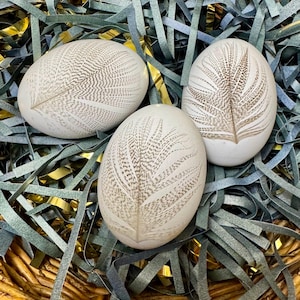 May include: Three white decorative eggs with a feather design in shades of brown, resting on a bed of blue and gold shredded paper. The eggs are nestled in a woven basket, creating a rustic and elegant display.