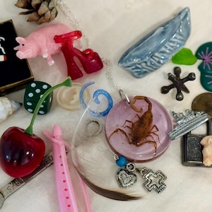 May include: A collection of assorted small objects including a pink pig figurine, a red cherry, a scorpion encased in a circular pink resin pendant, a metal wrench, a black square box with a cigarette graphic, a blue ceramic boat, and a yellow pin.