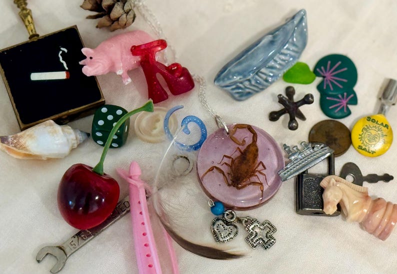 May include: A collection of assorted small objects including a pink pig figurine, a red cherry, a scorpion encased in a circular pink resin pendant, a metal wrench, a black square box with a cigarette graphic, a blue ceramic boat, and a yellow pin.