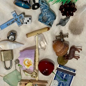 May include: A collection of miscellaneous small objects including a blue floral bell, a blue ceramic elephant, a light blue cross, a white die, a wooden pencil, a clear crystal, a brass padlock, a pink floral teacup, a red cherry, and a blue playing card.