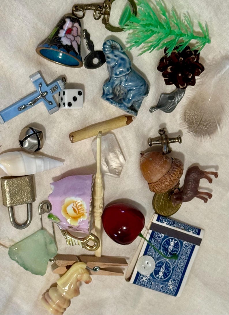 May include: A collection of miscellaneous small objects including a blue floral bell, a blue ceramic elephant, a light blue cross, a white die, a wooden pencil, a clear crystal, a brass padlock, a pink floral teacup, a red cherry, and a blue playing card.