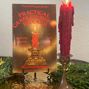Könnte beinhalten: Ein Buch mit dem Titel Practical Candleburning Rituals von Raymond Buckland mit einer roten Kerze auf dem Cover, auf einem schwarzen Metallständer neben einer hohen, brennenden, dunkelroten Stabkerze in einem bronzefarbenen Kerzenhalter.