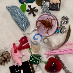 May include: A collection of assorted trinkets including a scorpion resin pendant, a pink pig figurine, a red cherry charm, a metal wrench, a seashell, a pinecone, a green die, a blue ceramic leaf, a metal padlock, and various small charms and tokens.