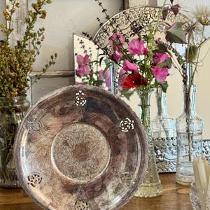 May include: A round silver-toned metal plate featuring an intricate embossed central pattern and six decorative cut-out sections along the scalloped rim. The plate is displayed upright on a black wire stand, showcasing a tarnished, aged metallic finish.