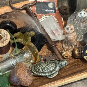 May include: A collection of assorted objects including a vintage metal key, a clear quartz crystal, a small ceramic turtle, a brown acorn, a miniature green plastic soldier, a wooden spool of thread, a glass salt shaker, and a small owl figurine.