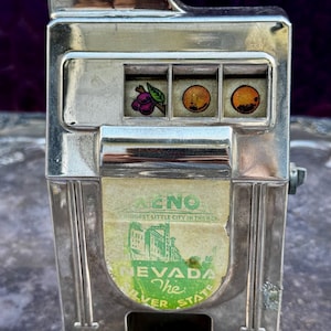 May include: A vintage silver-colored slot machine with three reels displaying fruit symbols. The top reads "10¢". The machine has a green and white label with the text "NEVADA The Silver State".