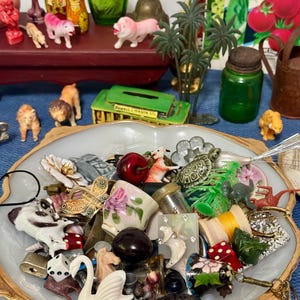 May include: A white scalloped bowl with gold trim filled with a collection of small trinkets including a white swan, a red cherry, a turtle, a miniature floral cup, dice, a spool of yellow thread, and various small animal and metal charms.