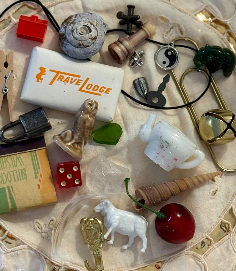 May include: A collection of miscellaneous small objects including a white Travelodge matchbox, a red die, a miniature white sheep figurine, a ceramic owl, a brass hook, a metal padlock, a shell, a glass teapot, a cherry, and a yin-yang charm.