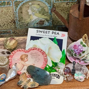 May include: A collection of vintage items including a rusty metal watering can, a Sweet Pea seed packet, a heart-shaped valentine card with a cherub, a miniature porcelain teacup and saucer with floral patterns, a spool of brown thread, and a butterfly.
