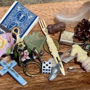 May include: A collection of small trinkets including a red cherry, a blue playing card, a floral porcelain piece, a wooden acorn, a blue crucifix, a plastic fork, a brown animal figure, a padlock, a pinecone, a green tree, and a chess piece.