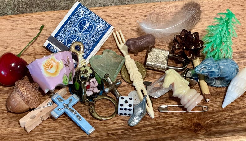 May include: A collection of small trinkets including a red cherry, a blue playing card, a floral porcelain piece, a wooden acorn, a blue crucifix, a plastic fork, a brown animal figure, a padlock, a pinecone, a green tree, and a chess piece.