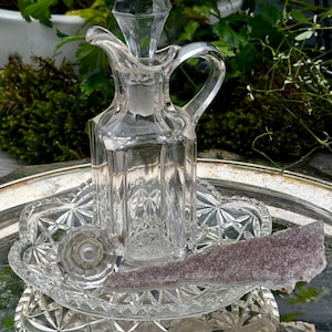 May include: A clear glass cruet with a diamond-shaped stopper and a handle, resting on a decorative glass dish. A piece of purple crystal sits on the dish. The items are displayed on a silver tray.