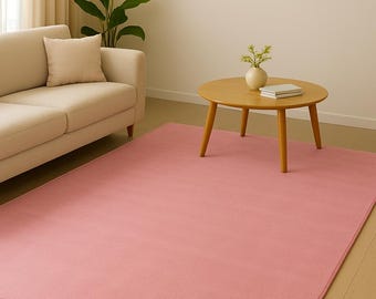 Op de afbeelding: Een woonkamer met een groot, rechthoekig, effen roze tapijt. Links staat een lichtbeige bank met een bijpassend kussen. In het midden staat een ronde houten salontafel met een vaas en boeken. Een ingelijst abstract kunstwerk hangt aan de muur.