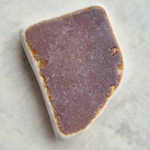 Op de afbeelding: Een close-up van een bruin en wit stuk zeeglas met een ruwe, onregelmatige vorm.