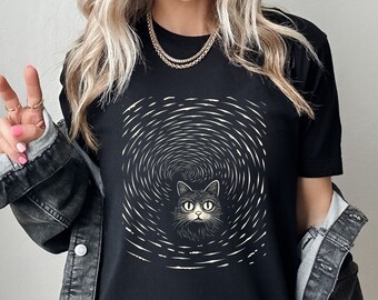 May include: A black short-sleeved t-shirt featuring a graphic print of a cat face centered within a white swirling vortex pattern. The design uses white lines to create a circular motion effect around the cat, which has large, wide eyes and detailed whiskers.