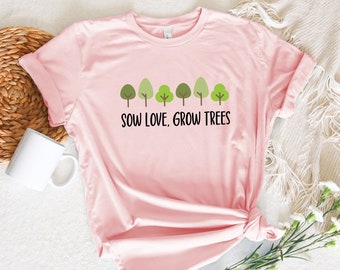 May include: A light pink short-sleeve t-shirt featuring a graphic of six stylized green trees in various shades and the black text "SOW LOVE, GROW TREES" printed across the center. The shirt has a crew neckline and a standard fit.