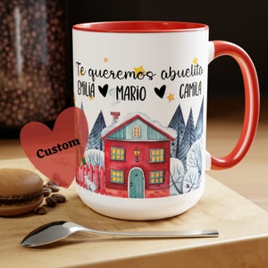 May include: White ceramic mug with a red rim and handle. The mug features a winter scene with a red house, snow-covered trees, and the text "Te queremos abuelita Emilia Mario Camila".