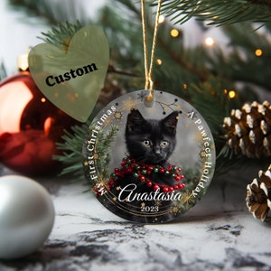 May include: A round Christmas ornament with a black kitten wearing a red and green beaded garland. The ornament has gold snowflakes and the text "My First Christmas" and "A Pawfect Holiday". The kitten's name, "Anastasia", and the year "2023" are also on the ornament.