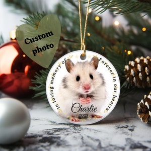 May include: A round, white ceramic ornament featuring a hamster photo, with the text "In loving memory of" and "Forever in our hearts." The hamster's name, "Charlie," and the year "2023" are also displayed. The ornament hangs from a gold string.